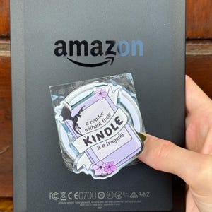 May include: A black Amazon Kindle e-reader with a rectangular acrylic pin attached to the back. The pin features a light purple book illustration with pink flowers, a black dragon silhouette, and the text: a reader without their KINDLE is a tragedy.