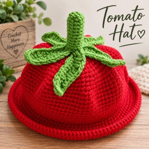 May include: A bright red crochet hat shaped like a tomato, featuring a green crocheted stem and leaves on top. The text Tomato Hat is printed in black script on the upper right side of the image, accompanied by a small heart symbol.