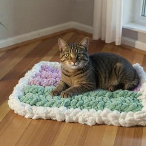 May include: A tabby cat rests on a handmade, rectangular pet bed. The bed features a pastel color scheme with sections of lavender, mint green, and white. The cat is brown and tan with green eyes. A potted plant is in the background.