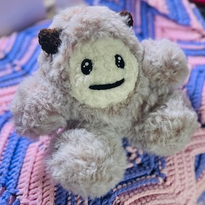 May include: A plush toy with a furry, light brown and white body. The toy has a white face with black eyes and a black smiling mouth. The toy is sitting on a pink and purple crocheted blanket.