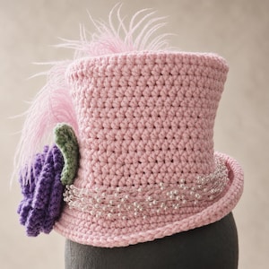 May include: A light pink crocheted top hat featuring a purple crocheted flower with a green leaf, a strand of white faux pearls wrapped around the base, and a tall, wispy light pink feather plume extending from the top of the hat.