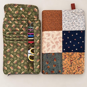 May include: A fabric organizer case with a green floral print interior featuring multiple slots holding various crochet hooks, safety pins, and scissors. The exterior is a patchwork design of six rectangular panels in brown, cream, and navy floral patterns.
