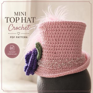 Puede incluir: Un mini sombrero de copa de ganchillo rosa con una flor de ganchillo morada, una hoja de ganchillo verde, un hilo de pequeñas cuentas blancas y una pluma rosa claro. El texto en la imagen dice MINI TOP HAT Crochet PDF PATTERN.