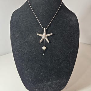 May include: A silver-toned necklace featuring a textured starfish pendant. A thin silver chain extends downward from the center of the starfish, ending in a small, round, off-white bead. The necklace is displayed on a black neck form.