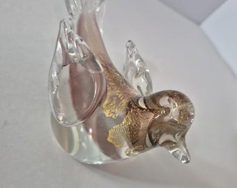 May include: A clear glass bird figurine featuring a smooth, rounded body with internal pinkish-purple hues and intricate gold-colored metallic flecks embedded within the glass along the back and head, with sculpted wings and tail.
