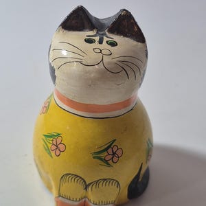 May include: A hand-painted ceramic cat figurine with a cream-colored face, black ears, and a yellow body decorated with small pink flowers and green leaves. The cat has a peach-colored collar and orange paws, with painted black whiskers and eyes.