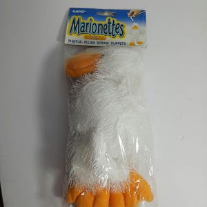 May include: A white plush duck puppet with orange feet and beak. The puppet is packaged in a clear plastic bag with a blue and white label that reads "Ganz Marionettes Duck Playful Plush String Puppets".
