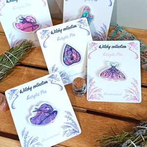 Op de afbeelding: Vijf heksenthema-acrylpinnen op witte kaartjes met lavendeltakjes en een klein flesje glitter. De pinnen tonen een heksenhoed, een spook, een mot, een kristal en een andere heksenhoed.