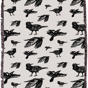 May include: A woven throw blanket featuring a repeating pattern of black birds on a cream background. The birds are shown in different poses, some with wings outstretched. The blanket has a fringed edge.