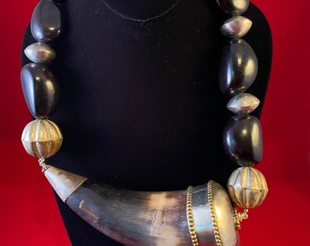 Peut inclure: Un collier imposant avec de grosses perles noires polies et des accents argentés. Il présente un pendentif en forme de corne avec des éléments métalliques argentés et une bordure dorée. Le collier est présenté sur un mannequin noir.