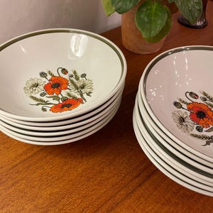 May include: A stack of white ceramic bowls with a floral design featuring orange poppies and white daisies. Each bowl has a green rim. The bowls are stacked on a wooden surface.