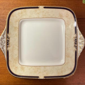 Puede incluir: Fuente cuadrada con centro blanco y borde color crema. El borde tiene un ribete azul marino y detalles dorados. La fuente tiene asas decorativas a cada lado.