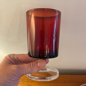 Könnte beinhalten: Ein rubinrotes Weinglas mit klarem Stiel und Fuß. Das Glas hat eine zylindrische Form und wird von einem klaren Stiel gehalten. Das Glas besteht aus einem transparenten Material.