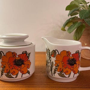 May include: White ceramic sugar bowl with lid and creamer featuring a floral design with orange poppies and wheat stalks. The set is on a wooden surface, with a plant in the background.