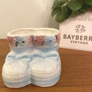 Op de afbeelding: Een vintage keramische babyschoenplanter in wit, blauw en roze. De planter heeft een strikdetail en een bloemmotief rond de bovenkant. De woorden "Curated by BAYBERRY VINTAGE" zijn zichtbaar op de achtergrond.