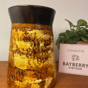 Könnte beinhalten: Eine Vintage-Keramikvase mit strukturierter Oberfläche. Die Vase hat einen dunkelbraunen Rand und einen Körper mit gelben, orangefarbenen und braunen Farbtönen. Die Vase ist mit "Curated by BAYBERRY VINTAGE" beschriftet.