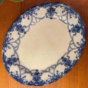 May include: Oval antique plate with a white centre and a blue floral border. The blue design features intricate scrollwork and decorative elements. The plate has a scalloped edge, likely made of porcelain or ceramic.