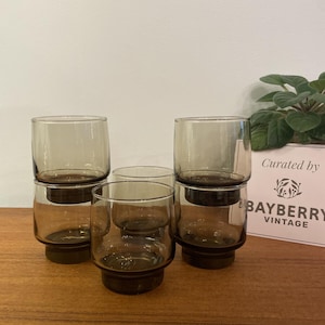 May include: A collection of six vintage, stackable, smoked glass tumblers. The glasses feature a cylindrical design with a dark brown base and a lighter, smoky grey upper section. The glasses are displayed on a wooden surface.