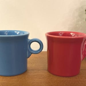 May include: Two ceramic mugs with a smooth, glossy finish. One mug is a solid medium blue, and the other is a solid bright red. Both mugs feature a cylindrical body with a slightly flared rim and a small, circular handle attached to the side.