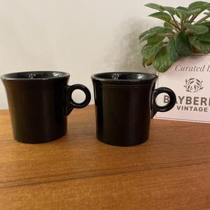 Könnte beinhalten: Zwei schwarze Keramikbecher mit abgerundeten Henkeln stehen auf einer Holzoberfläche. Die Becher haben ein schlichtes Design. Eine kleine Pflanze und ein Schild mit der Aufschrift "Curated By Bayberry Vintage" sind im Hintergrund.