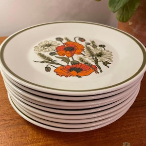 May include: A stack of white ceramic plates with a floral design featuring orange poppies and other flowers. Each plate has a green border. The plates are stacked on a wooden surface.