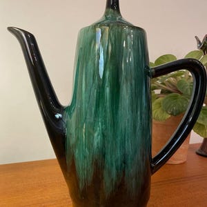 Puede incluir: Una cafetera alta de cerámica con acabado brillante. La cafetera presenta una combinación de colores verde oscuro y negro, con un pico largo y un asa curvada. La tapa está rematada con un pequeño pomo redondeado. Probablemente vintage o retro.