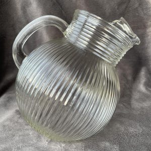 Puede incluir: Jarra de vidrio transparente con un diseño diagonal acanalado. La jarra tiene un asa curva y un pico para verter. El vidrio transparente permite ver el contenido. Una pieza clásica para servir bebidas.