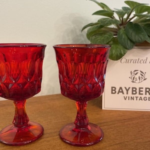May include: Two vintage ruby red wine glasses with a textured diamond pattern. The stemware is displayed on a wooden surface, with a plant and a sign that reads "BAYBERRY VINTAGE" in the background.