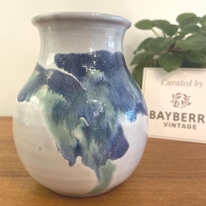 May include: A ceramic vase with a rounded body and a flared rim. The vase is mainly white with blue and green abstract paint strokes. The text "Curated by BAYBERRY VINTAGE" is on a white card in the background.