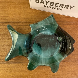 Puede incluir: Un plato de cerámica con forma de pez que presenta un esmalte brillante en tonos verde bosque profundo y azul verdoso con un centro oscuro. El plato tiene un pequeño detalle de ojo circular en relieve. Una tarjeta blanca con el texto BAYBERRY VINTAGE es visible en la parte superior.