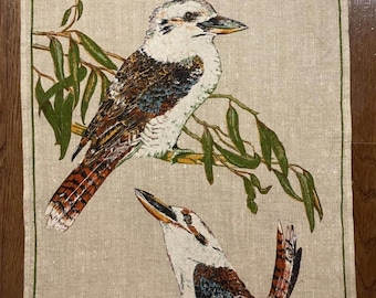 May include: Linen tea towel featuring two kookaburras perched on branches with green leaves. The birds have white, brown, and blue plumage. The text "Kookaburras Australia" is at the top. The towel has a green border.