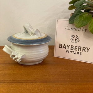 May include: A ceramic lidded bowl with a white body and a blue rim. The lid has a handle and a yellow accent. The bowl is on a wooden surface. A sign that says "Curated by BAYBERRY VINTAGE" is in the background.