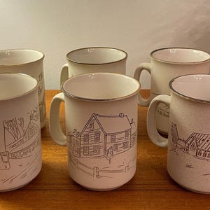 Puede incluir: Juego de seis tazas de cerámica blanquecina con borde y asa marrones. Cada taza presenta un dibujo lineal negro diferente de una escena de paisaje, incluyendo una casa, una iglesia y una escena invernal. Las tazas están sobre una superficie de madera.