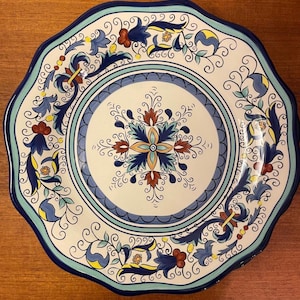 May include: A decorative ceramic plate with a scalloped edge. The plate features a white background with a blue, yellow, and red floral design. The rim is a dark blue, and the plate has a diameter of approximately 30 cm.
