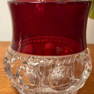 Puede incluir: Un cuenco de vidrio vintage con una sección superior de color rojo rubí y una sección inferior transparente con dibujos. La sección transparente presenta diseños circulares repetidos con patrones en forma de estrella. El cuenco está sobre una superficie de madera.