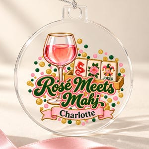 Op de afbeelding: Een doorzichtig, rond, plat ornament met een afbeelding van een wijnglas gevuld met roze vloeistof, drie mahjongtegels en de tekst "Ros&eacute; Meets Mahj" boven een roze banner met de naam "Charlotte" in het zwart, omringd door gekleurde stippen.