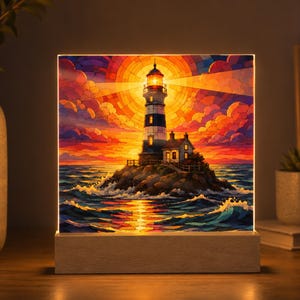 K&ouml;nnte beinhalten: Ein quadratischer, beleuchteter Leuchtkasten mit einer lebendigen Illustration im Buntglasstil, die einen Leuchtturm auf einer felsigen Insel zeigt. Die Szene stellt einen Sonnenuntergang mit orangefarbenen, roten und violetten Wolken dar.