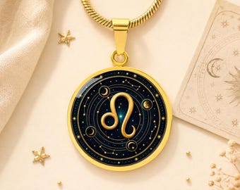 May include: A gold-toned circular pendant necklace featuring the Leo zodiac symbol in the centre. The design includes a dark blue background with gold celestial elements, including stars, constellations, and moon phases, encased in a polished gold frame.