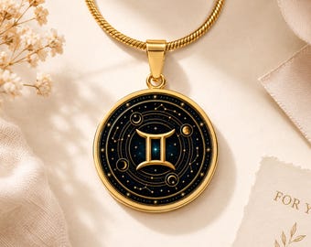 May include: A gold-toned circular pendant necklace featuring the Gemini zodiac symbol in the centre. The design includes a dark blue background with gold celestial elements, stars, and moon phases, all encased in a polished gold metal frame.