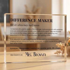 May include: A clear acrylic block with the words "DIFFERENCE MAKER" and a definition, synonyms, and "Mrs. Brown" printed on it. The block is on a wooden surface with flowers in the background.