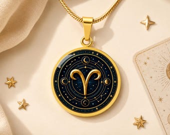 May include: A gold-toned circular pendant necklace featuring the Aries zodiac symbol in the centre. The design includes a dark blue background with celestial motifs, including stars, constellations, and moon phases, all encased in a polished gold frame.