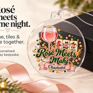 Op de afbeelding: Een doorzichtig, rond, plat ornament met een afbeelding van een wijnglas gevuld met roze vloeistof, drie mahjongtegels en de tekst "Ros&eacute; Meets Mahj" boven een roze banner met de naam "Charlotte" in het zwart, geaccentueerd door kleurrijke stippen.
