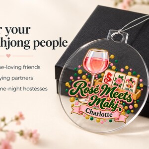 Op de afbeelding: Een rond ornament van helder acryl met een afbeelding van een wijnglas gevuld met roze vloeistof, drie mahjongtegels en de tekst "Ros&eacute; Meets Mahj" met de naam "Charlotte" op een roze lint, geaccentueerd door kleine groene en gouden stippen.