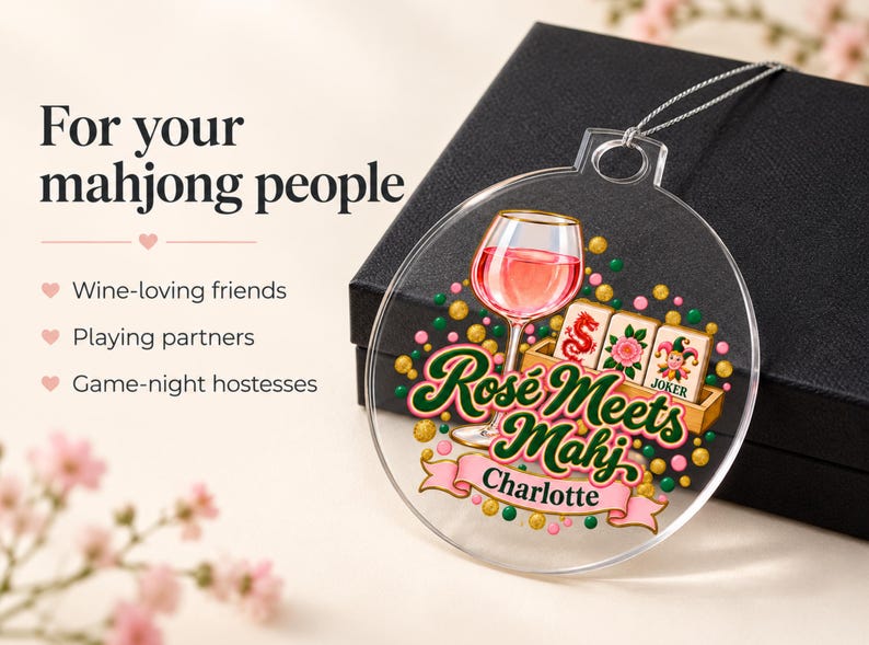 Op de afbeelding: Een rond ornament van helder acryl met een afbeelding van een wijnglas gevuld met roze vloeistof, drie mahjongtegels en de tekst "Ros&eacute; Meets Mahj" met de naam "Charlotte" op een roze lint, geaccentueerd door kleine groene en gouden stippen.