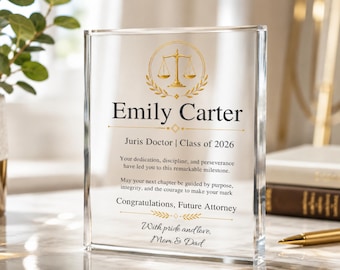 May include: A clear acrylic plaque with gold detailing, inscribed with "Emily Carter" and "Juris Doctor | Class of 2026." The plaque features a scales of justice emblem and a message of congratulations from "Mom & Dad."