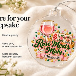 Op de afbeelding: Een ronde, transparante acryl hanger met een afbeelding van een wijnglas met roze vloeistof, drie mahjongtegels en de tekst "Ros&eacute; Meets Mahj" boven een roze banner met de naam "Charlotte" in zwarte letters, hangend aan een zilverkleurig koord.