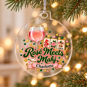 Op de afbeelding: Een doorzichtig, rond ornament van acryl met een afbeelding van een wijnglas gevuld met roze vloeistof, drie mahjongtegels en de tekst "Ros&eacute; Meets Mahj" boven een roze banner met de naam "Charlotte" in het zwart, omringd door kleurrijke stippen.