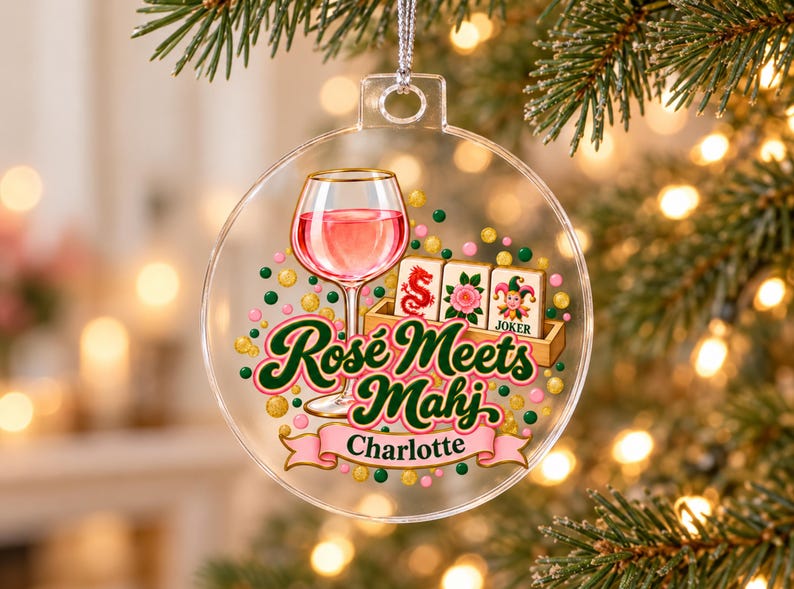 Op de afbeelding: Een doorzichtig, rond ornament van acryl met een afbeelding van een wijnglas gevuld met roze vloeistof, drie mahjongtegels en de tekst "Ros&eacute; Meets Mahj" boven een roze banner met de naam "Charlotte" in het zwart, omringd door kleurrijke stippen.