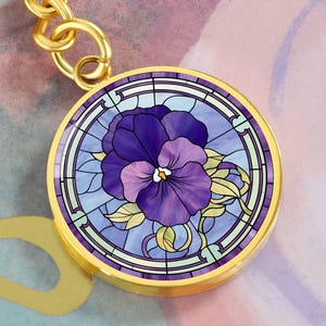 May include: A gold-toned keychain featuring a round charm with a stained glass design of a purple pansy flower.