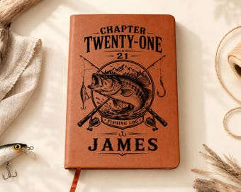 May include: A tan leather-bound journal featuring a dark brown engraved design of a fish, two crossed fishing rods, and mountains. The text reads CHAPTER TWENTY-ONE, 21, FISHING LOG, and JAMES. A ribbon bookmark extends from the bottom edge.